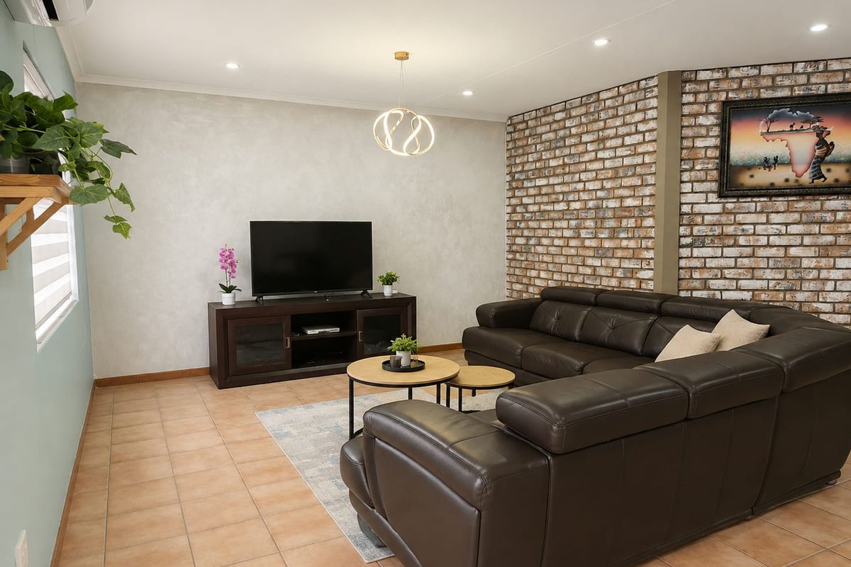A well-maintained living room featuring a distinctive exposed brick feature wall, neutral tiled flooring, and modern recessed lighting.