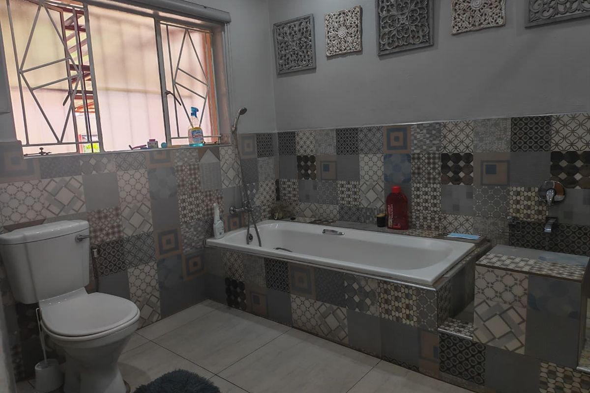 A bathroom featuring a built-in bathtub, toilet, and decorative patterned wall tiles with security bars on the window.