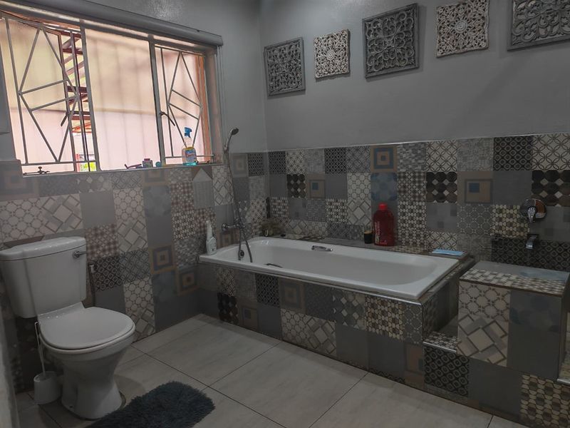 A bathroom featuring a built-in bathtub, toilet, and decorative patterned wall tiles with security bars on the window.