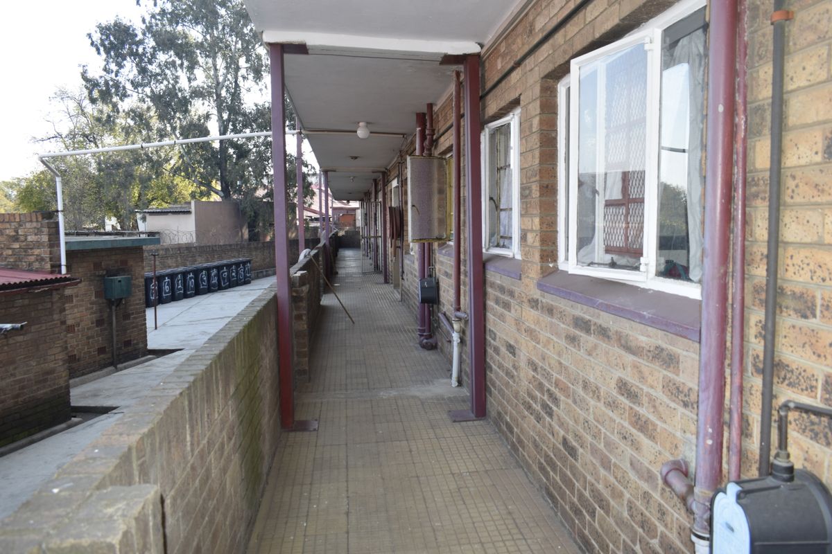 An exterior walkway featuring brick walls, metal support columns, and security window grilles, providing access to apartment units.
