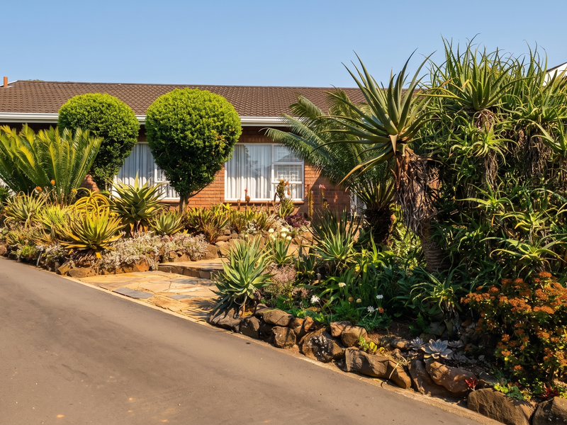 A well-maintained single-story brick house featuring a tiled roof, asphalt driveway, and established landscaped garden with stone retaining walls.