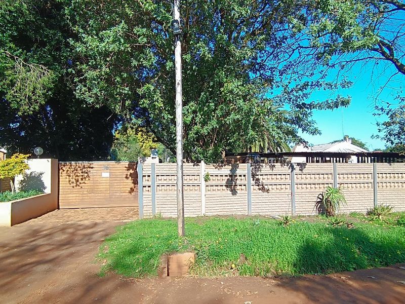 Front view of the property featuring a secure brick boundary wall, wooden driveway gate, and established garden area with mature trees.