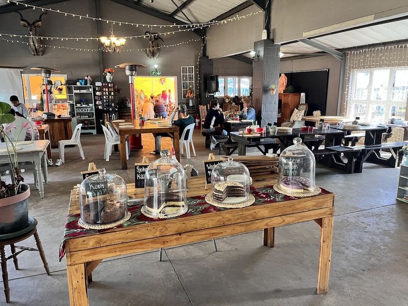 Spacious open-plan interior featuring exposed steel roof trusses, concrete flooring, and large windows, currently utilized as a rustic dining and cafe area.
