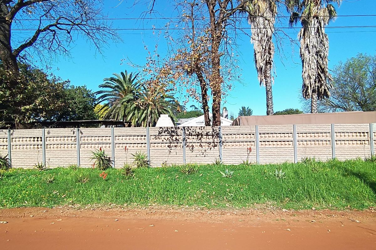 Street view of the property showing a precast concrete boundary wall bordering a dirt road with mature trees in the background.