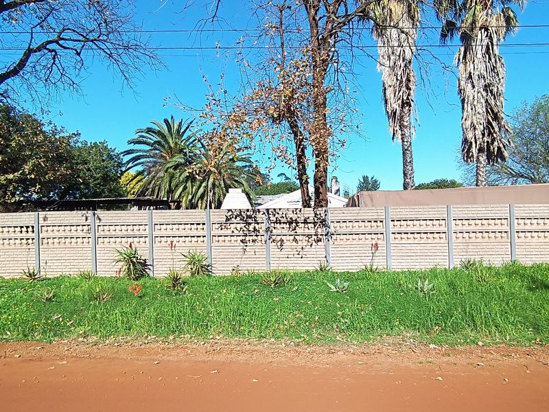 Street view of the property showing a precast concrete boundary wall bordering a dirt road with mature trees in the background.