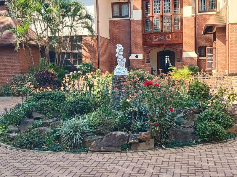 A well-maintained front exterior featuring a brick paved driveway, landscaped stone garden bed, and a distinctive architectural facade with leaded glass windows.