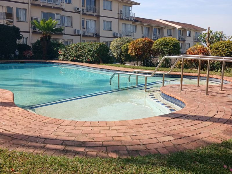 A communal swimming pool area featuring brick paving, metal handrails, and landscaped garden surrounds set against an apartment building facade.