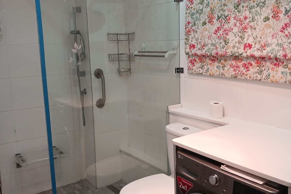 A functional bathroom featuring a glass-enclosed shower with a rain shower head, a toilet, and an integrated laundry appliance with a clean, modern finish.