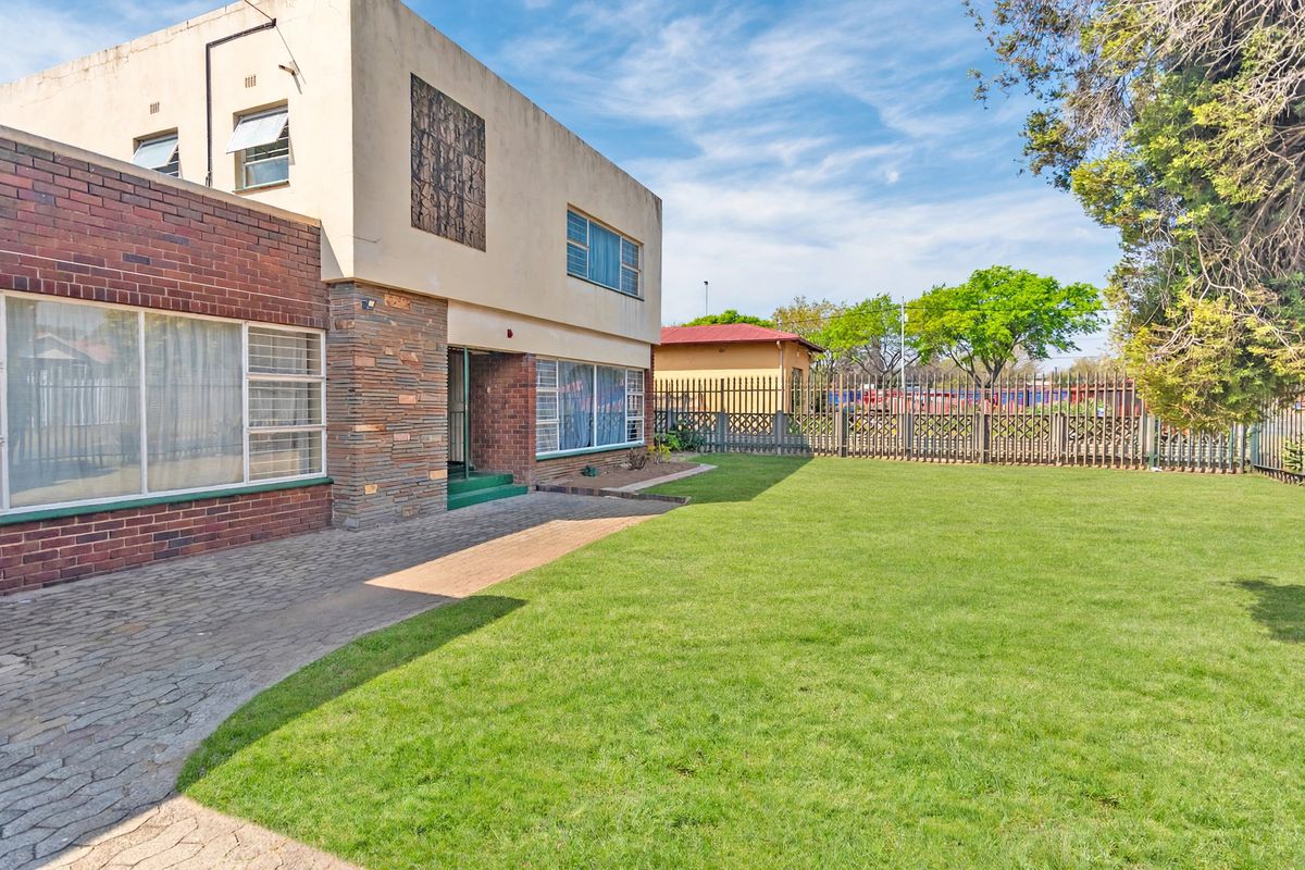 Rear view of a two-story brick and plaster house featuring a paved patio area, a spacious lawn, and secure metal perimeter fencing.