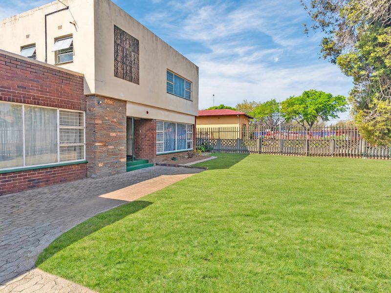 Rear view of a two-story brick and plaster house featuring a paved patio area, a spacious lawn, and secure metal perimeter fencing.