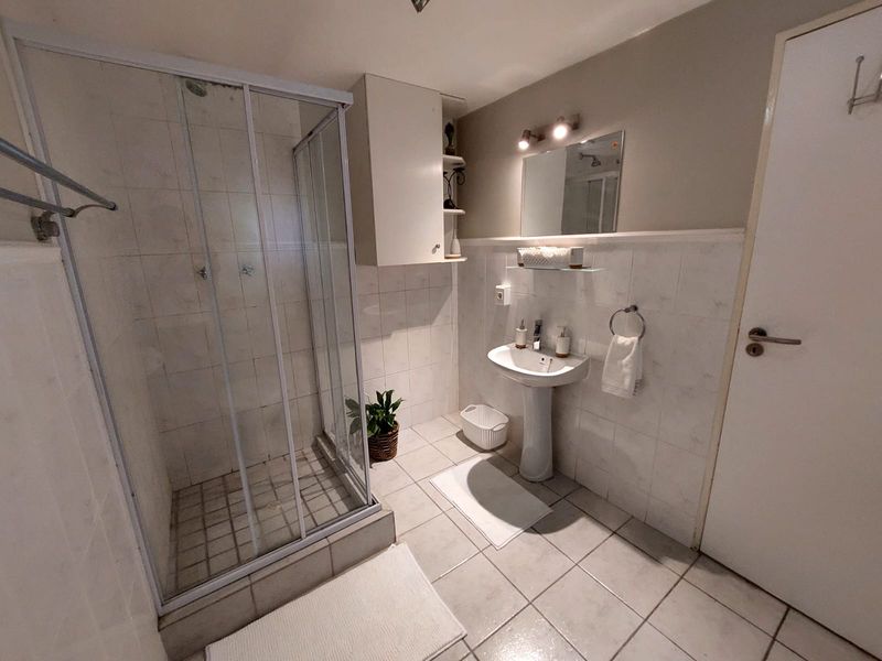 A functional bathroom featuring a glass shower enclosure, pedestal sink, wall-mounted storage cabinet, and neutral tiled finishes throughout.