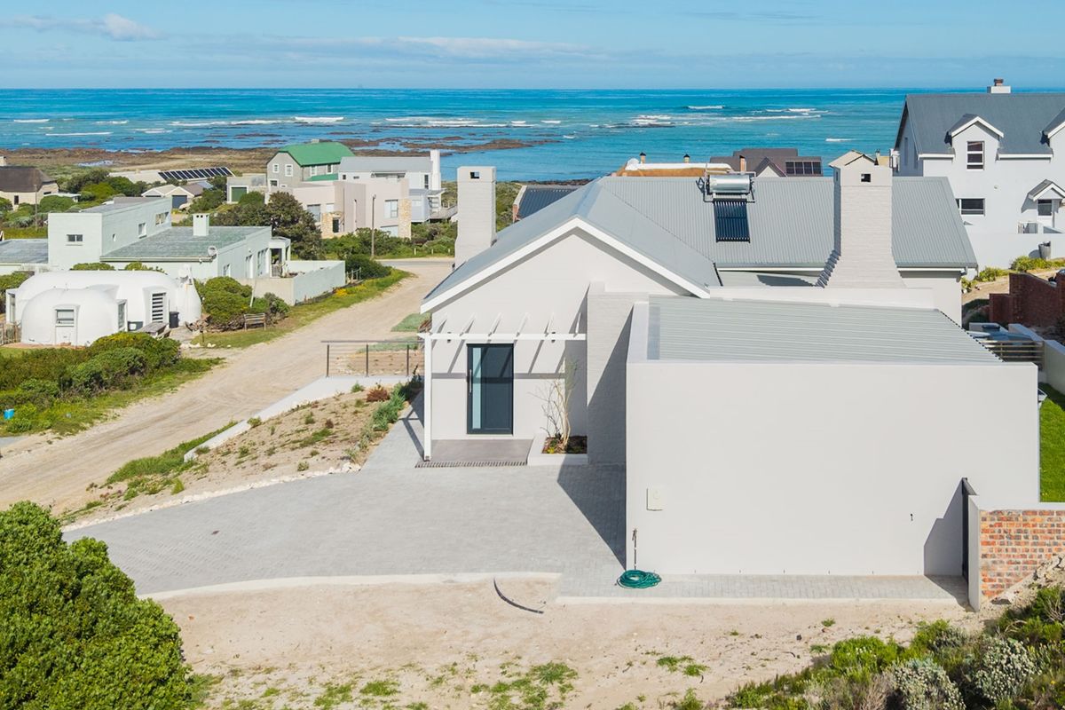 Aerial view of a modern coastal home featuring a metal roof, solar water heating system, and a paved driveway, set in a scenic seaside location.