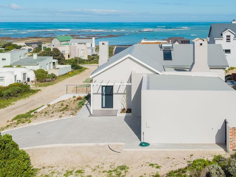 Aerial view of a modern coastal home featuring a metal roof, solar water heating system, and a paved driveway, set in a scenic seaside location.