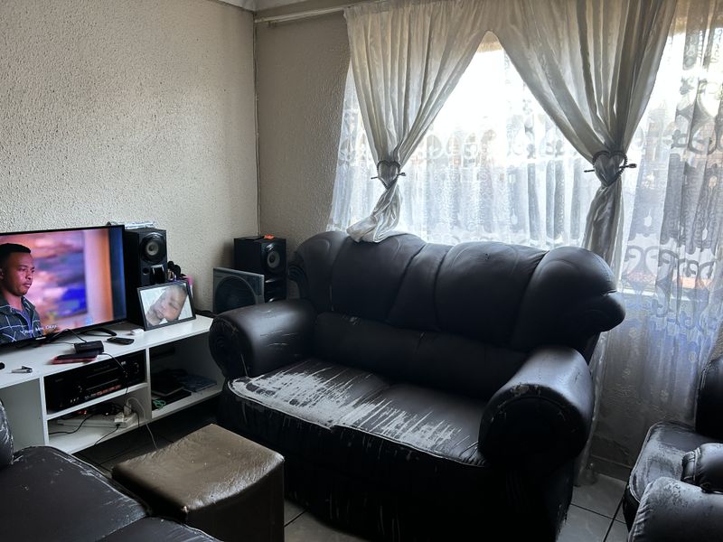 A living room area featuring tiled flooring and window treatments, currently showing signs of wear on the wall finishes.