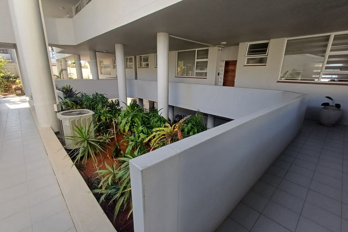 An open-air apartment complex walkway featuring tiled flooring, structural concrete columns, and a central landscaped garden area.