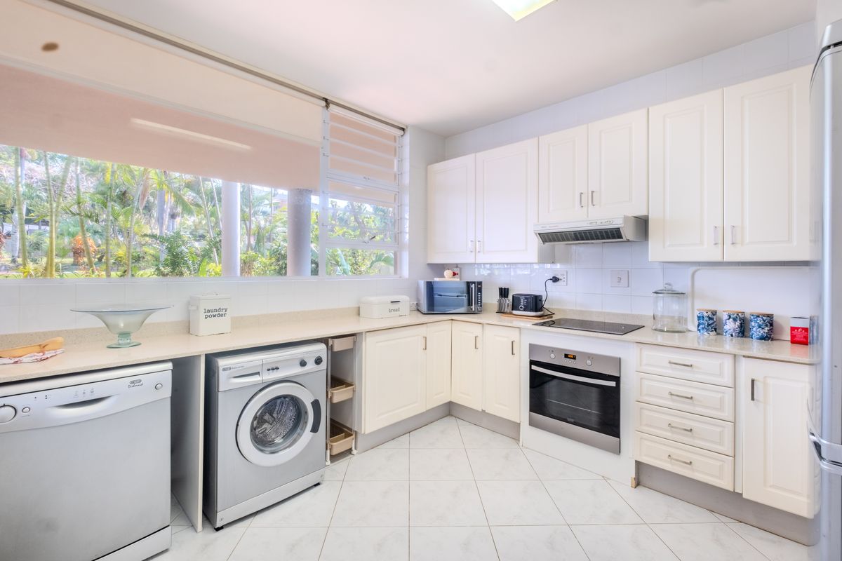 A functional kitchen featuring white cabinetry, stone countertops, integrated oven and hob, and tiled flooring with large windows overlooking a garden.