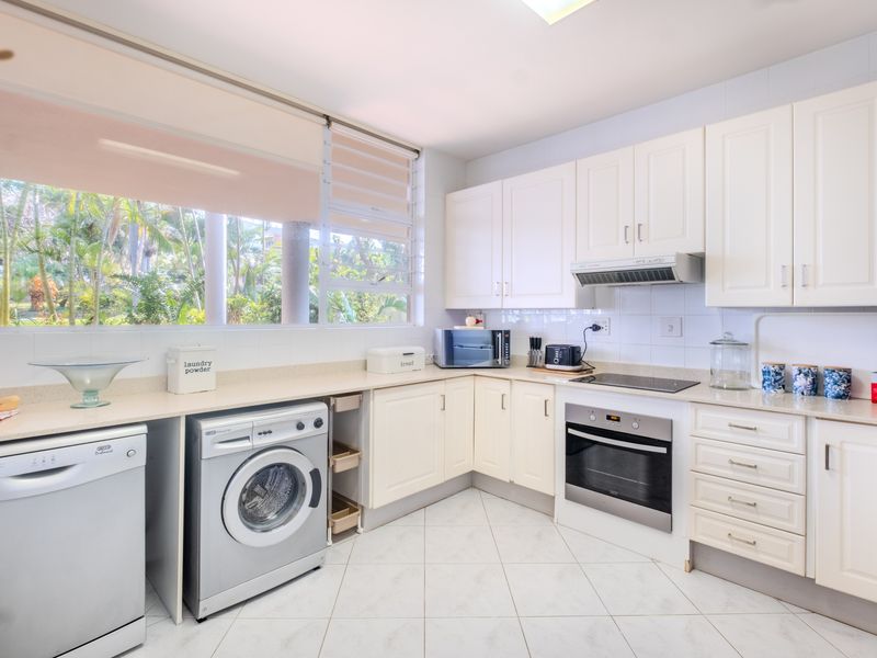 A functional kitchen featuring white cabinetry, stone countertops, integrated oven and hob, and tiled flooring with large windows overlooking a garden.