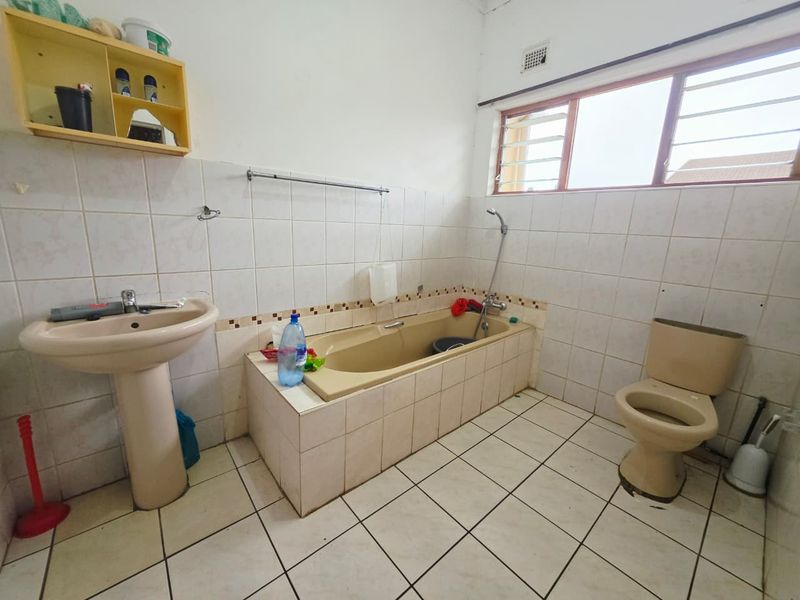 A dated bathroom featuring a pedestal basin, built-in bathtub, and toilet with tiled finishes and a window providing natural light.