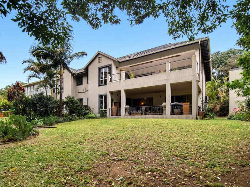 Rear view of the apartment building featuring a spacious lawn, covered ground-floor patio, and an upper-level balcony with metal railings.