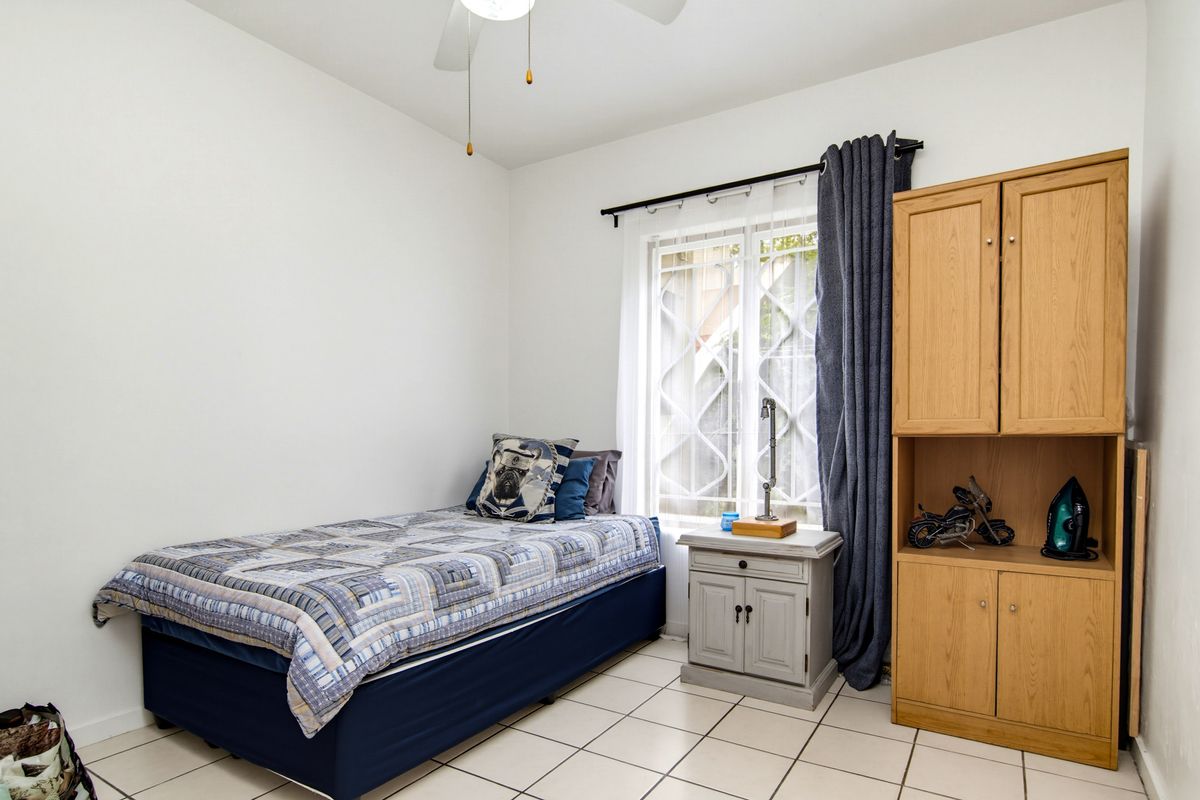 A functional bedroom featuring tiled flooring, a ceiling fan, and a window fitted with security bars, providing a clean and practical living space.