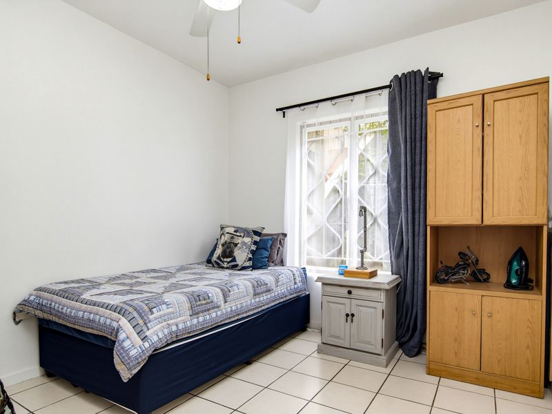 A functional bedroom featuring tiled flooring, a ceiling fan, and a window fitted with security bars, providing a clean and practical living space.
