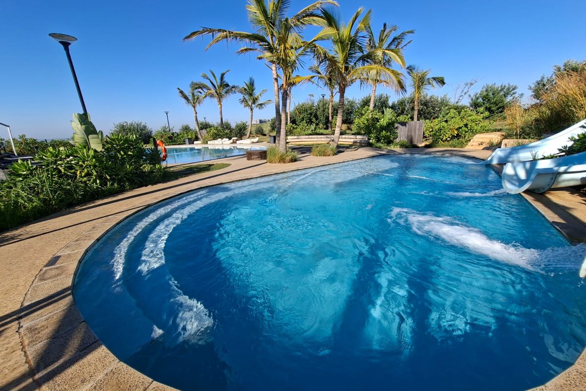 A well-maintained outdoor swimming pool area featuring a built-in water slide, paved decking, and established palm trees in a landscaped setting.