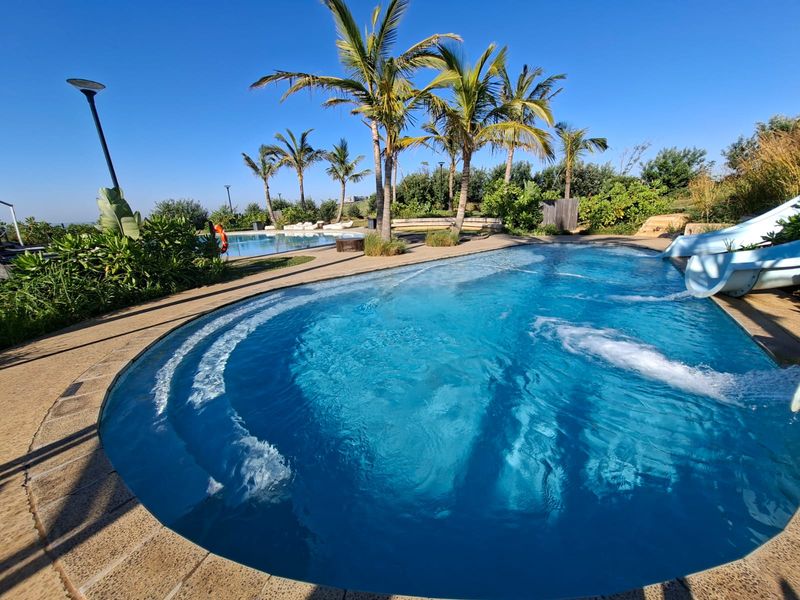 A well-maintained outdoor swimming pool area featuring a built-in water slide, paved decking, and established palm trees in a landscaped setting.