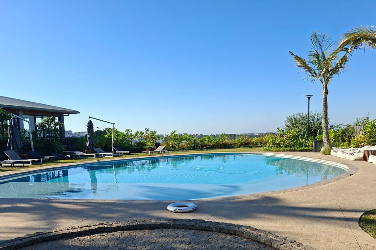 A well-maintained outdoor swimming pool area featuring a paved deck, landscaped garden surroundings, and mature palm trees under a clear blue sky.