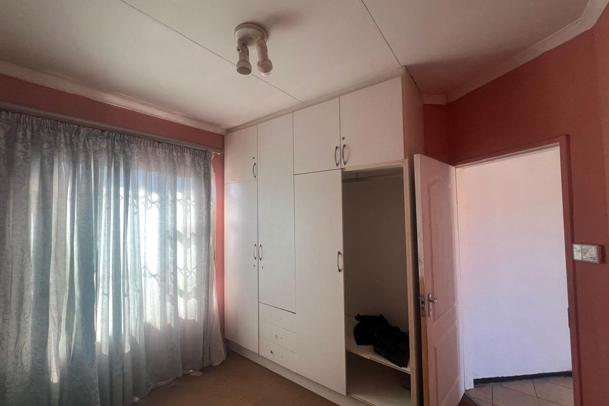 A bedroom featuring built-in white cupboards, carpeted flooring, and a window with security bars, presenting a blank canvas for renovation.