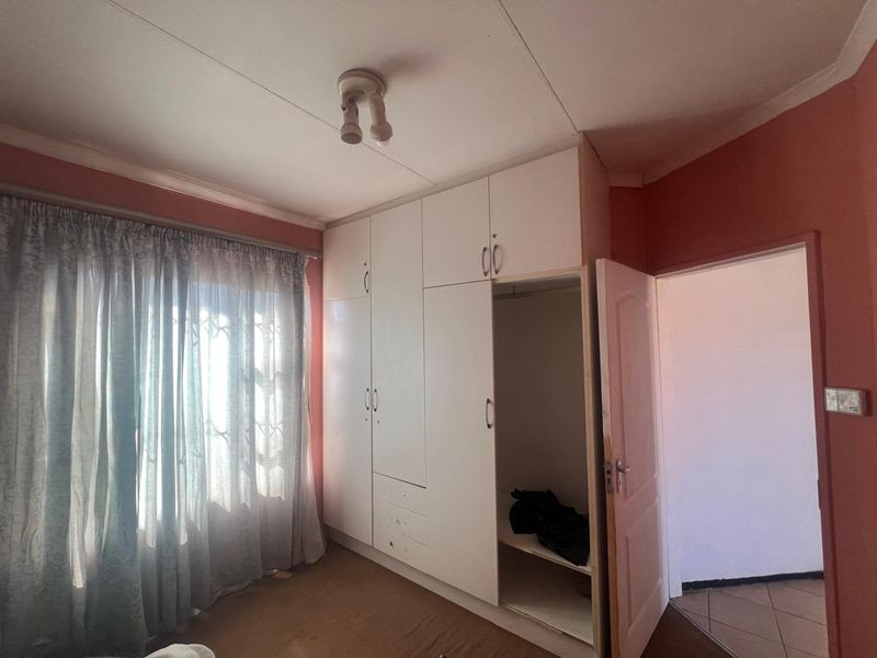 A bedroom featuring built-in white cupboards, carpeted flooring, and a window with security bars, presenting a blank canvas for renovation.