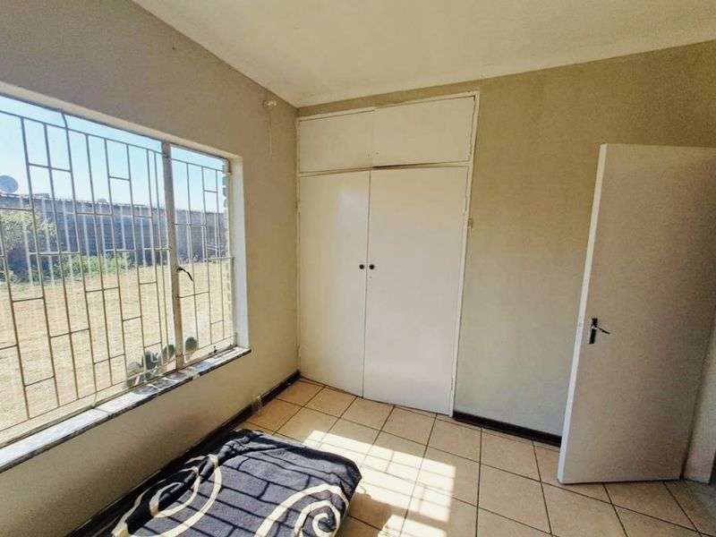 A simple bedroom featuring tiled flooring, built-in cupboards, and large windows with security bars, offering a functional space ready for updates.