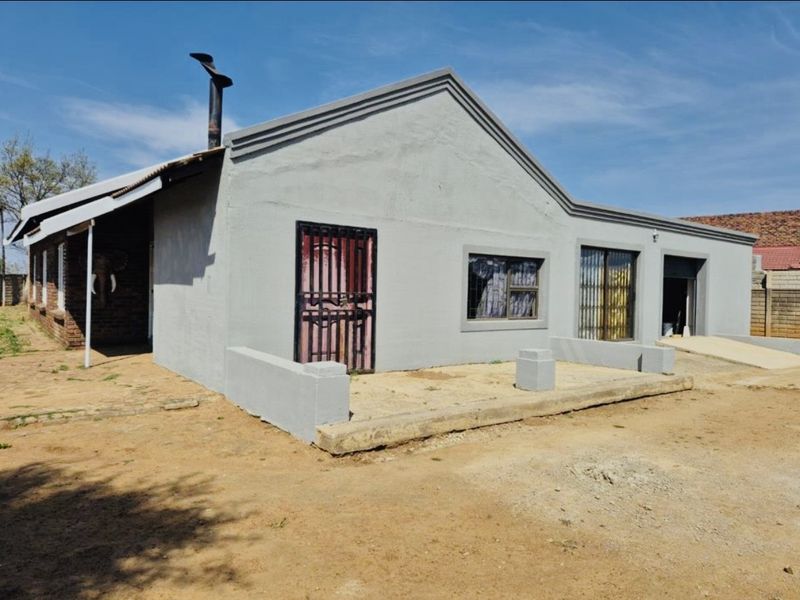 Front exterior view of a single-story house featuring a concrete porch, metal security gates, and a chimney flue, set on an unpaved lot.