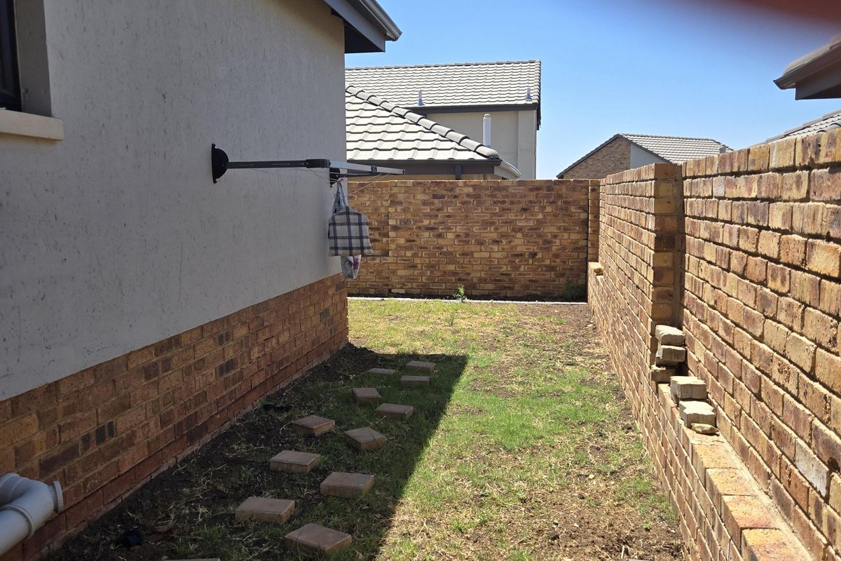 A narrow side yard featuring a brick boundary wall, paved stepping stones, and a wall-mounted clothesline, requiring landscaping and maintenance.