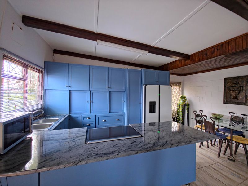 Open-plan kitchen and dining area featuring blue cabinetry, granite countertops, an integrated hob, and exposed timber ceiling beams.
