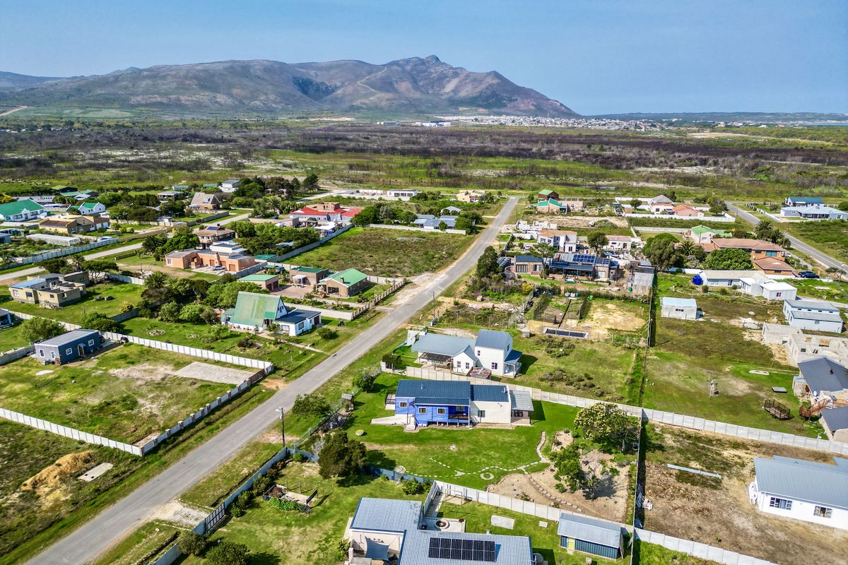 Aerial view of a residential neighborhood featuring houses with metal roofs, solar panels, and surrounding garden plots with mountain views in the distance.