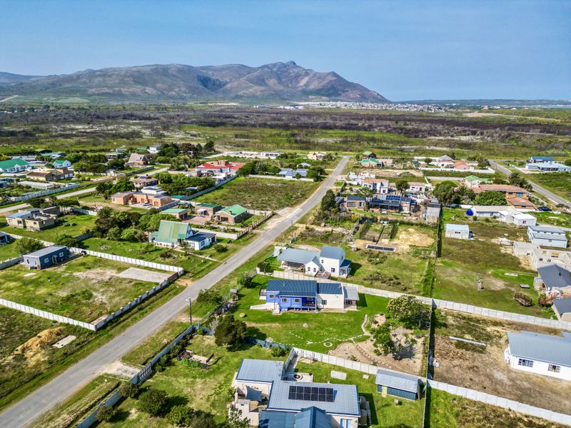 Aerial view of a residential neighborhood featuring houses with metal roofs, solar panels, and surrounding garden plots with mountain views in the distance.