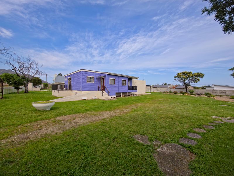 Rear view of a detached house featuring a raised timber deck, concrete access ramp, and corrugated metal siding set within a large garden area.