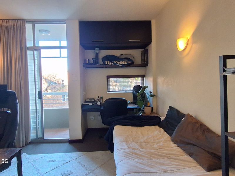 A functional bedroom featuring a built-in desk with overhead storage, wall-mounted lighting, and direct access to a private balcony.