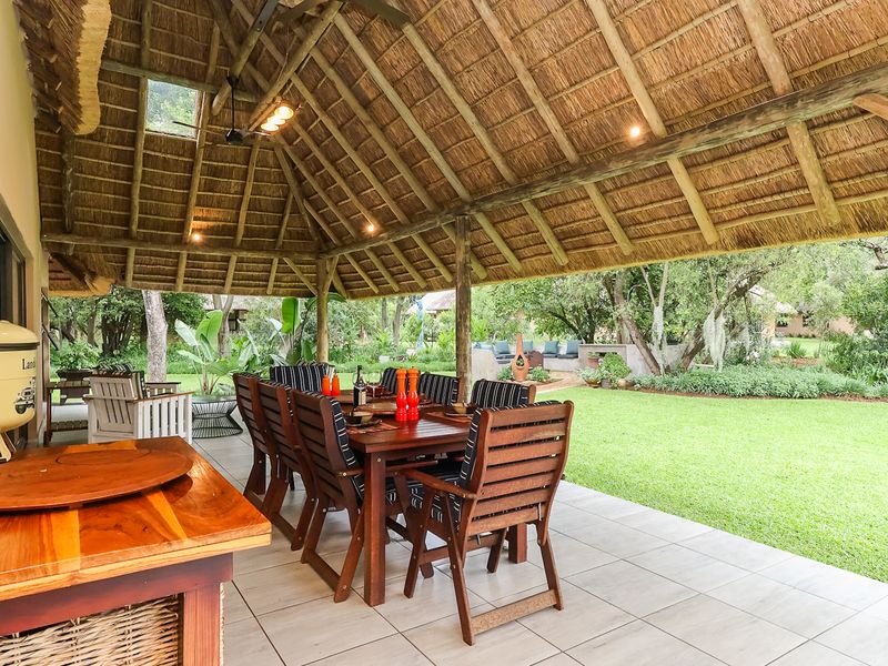 A spacious covered patio featuring a traditional thatched roof with exposed timber beams, tiled flooring, and direct access to a lush garden.