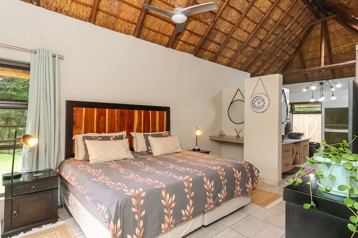 Spacious bedroom featuring a high thatched roof with exposed timber rafters, ceiling fan, tiled flooring, and an integrated en-suite bathroom area.