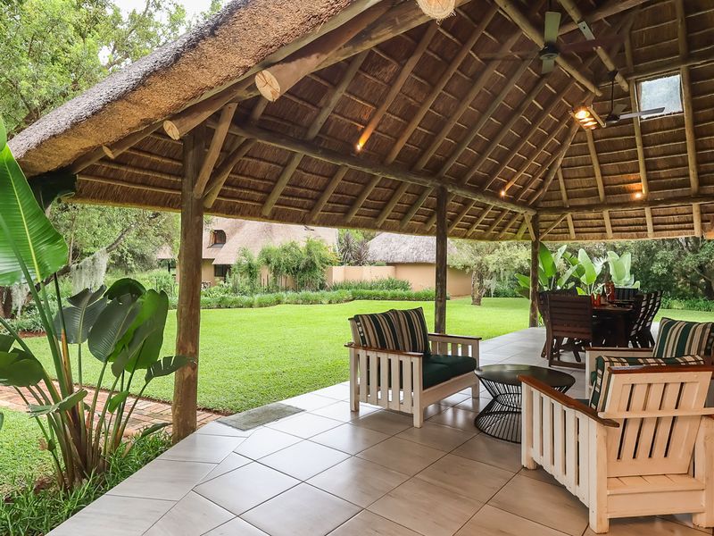 A spacious covered patio featuring a traditional thatched roof, timber support beams, and tiled flooring, overlooking a well-maintained garden.