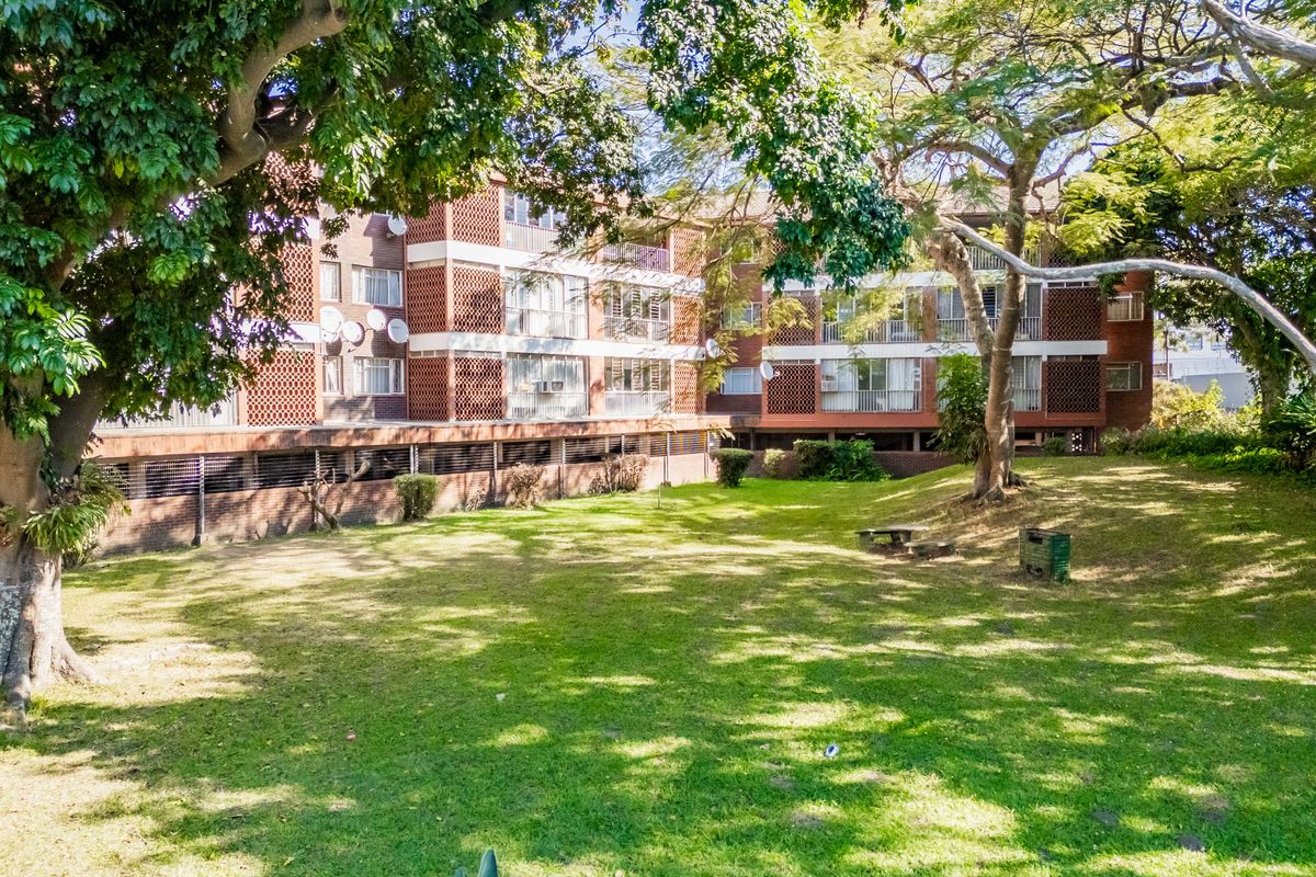 A view of the rear apartment building facade featuring brick architecture, private balconies, and a well-maintained communal garden area with mature trees.
