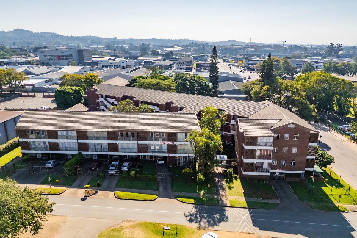 Aerial view of the Pinefern apartment complex, showcasing the multi-story brick building, tiled roofing, and surrounding paved parking areas and landscaping.