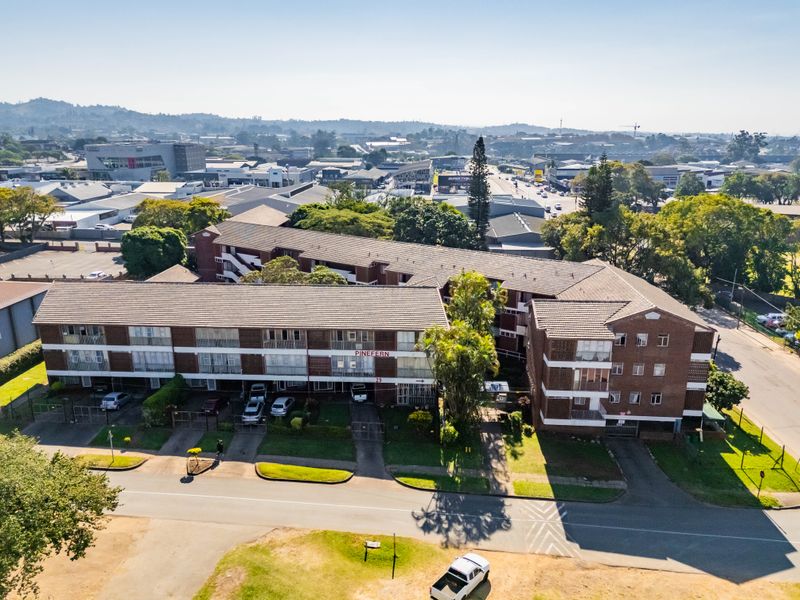 Aerial view of the Pinefern apartment complex, showcasing the multi-story brick building, tiled roofing, and surrounding paved parking areas and landscaping.