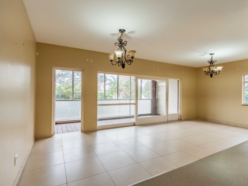 Spacious, empty living area featuring neutral tiled flooring, dual wrought iron chandeliers, and large sliding glass doors providing access to an attached balcony.