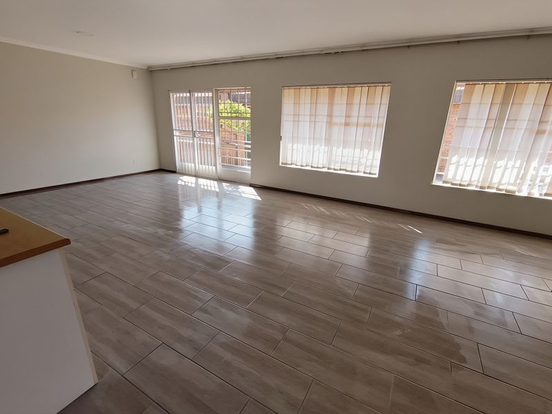 A spacious, empty living area featuring modern wood-look tiled flooring, large windows with vertical blinds, and a secure glass door leading outdoors.