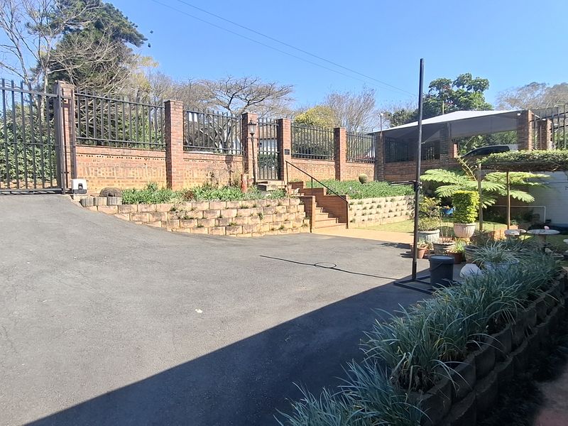 Front exterior view featuring an asphalt driveway, brick retaining walls, metal security fencing, and a paved walkway leading to the property entrance.