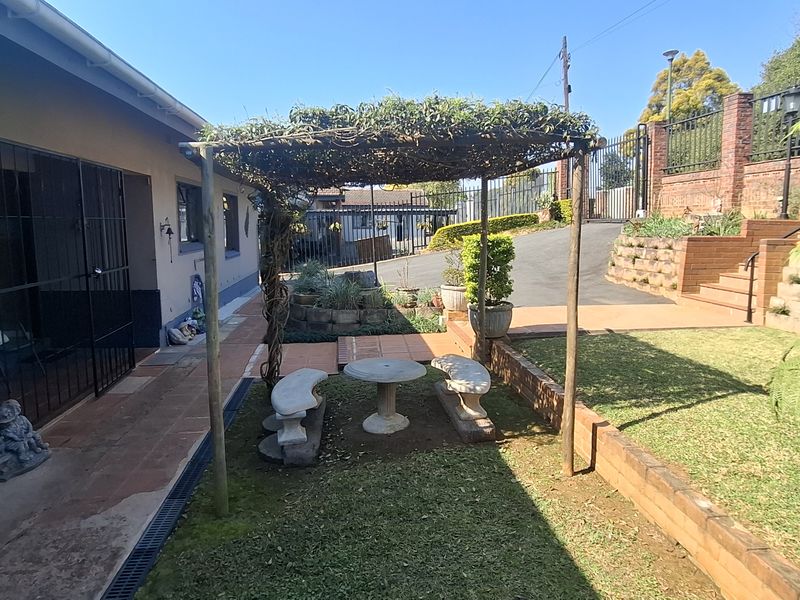 Rear garden area featuring a paved driveway, brick retaining walls, and a shaded pergola structure set within a landscaped lawn.