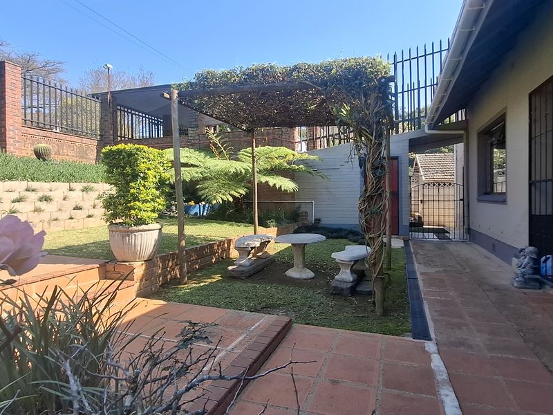 A private rear garden featuring a terracotta paved patio, a pergola-covered seating area, and tiered retaining walls with established landscaping and security fencing.