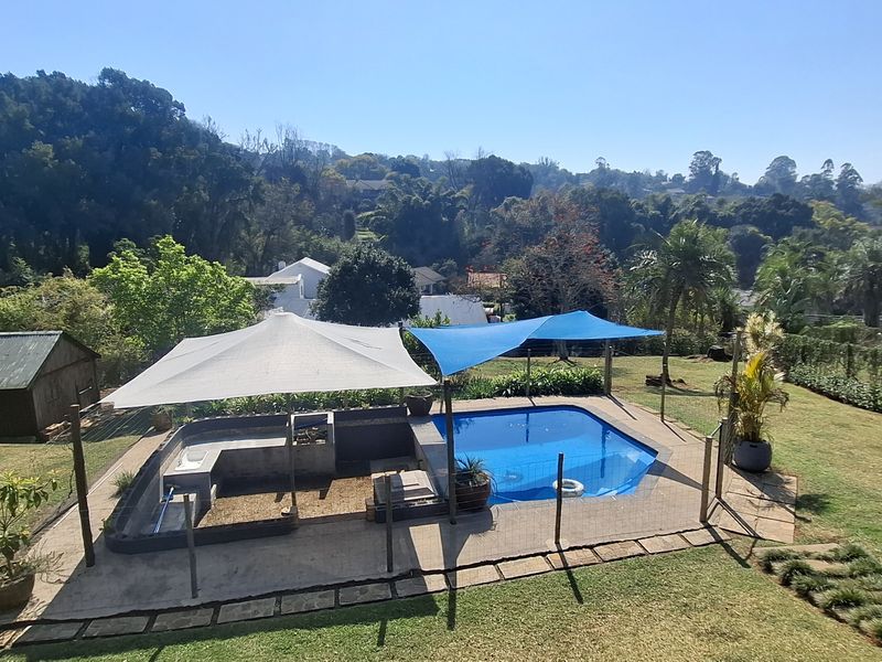 Outdoor pool area featuring a swimming pool, built-in braai, and shade sails, set within a landscaped garden with scenic views.